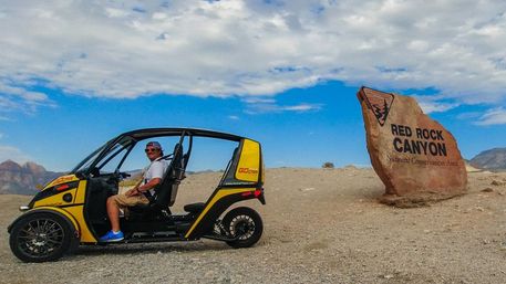 Person in a yellow open-air tour vehicle parked beside a large sandstone sign for Red Rock Canyon National Conservation Area, arid desert landscape and blue cloudy sky