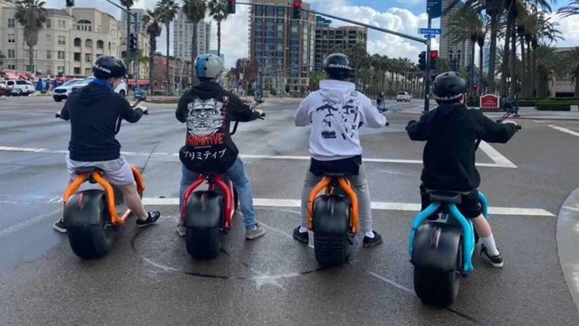 Four helmeted riders on colorful electric minibikes (orange, red, blue) stopped at a wet city intersection with palm trees, high-rise buildings, and traffic lights overhead, ready to roll.