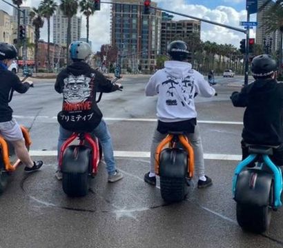 Four helmeted riders on colorful electric minibikes (orange, red, blue) stopped at a wet city intersection with palm trees, high-rise buildings, and traffic lights overhead, ready to roll.