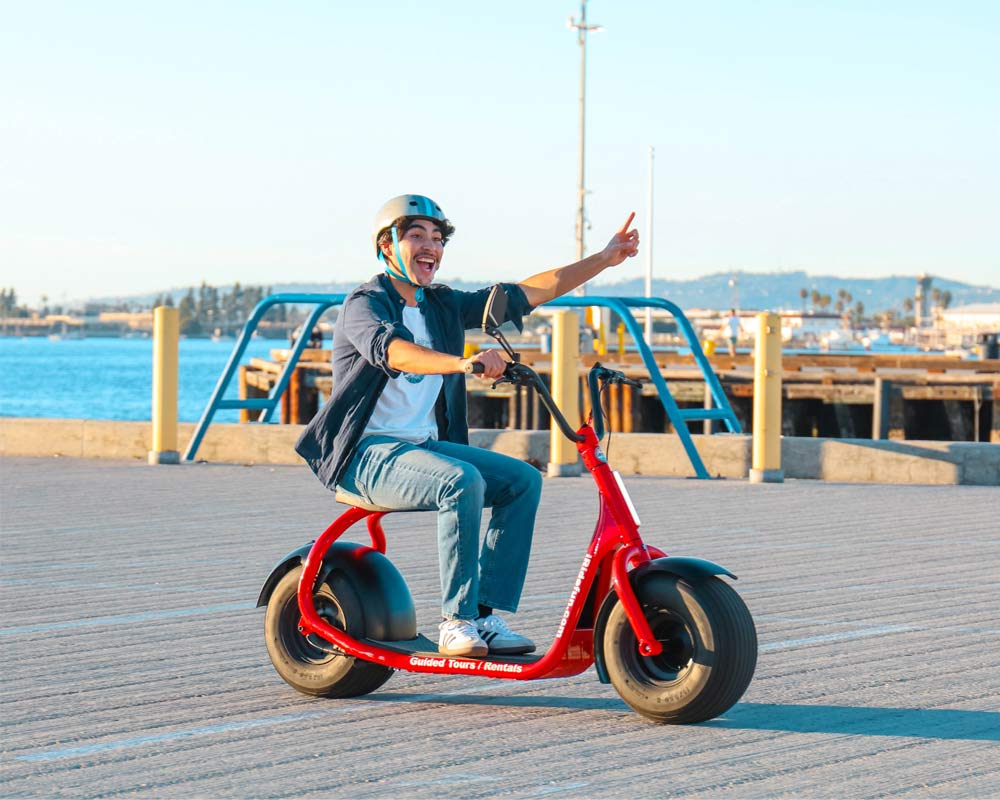 Smiling rider in a helmet on a red fat-tire electric scooter cruising a sunny waterfront pier/boardwalk, pointing toward the bay and distant hills.