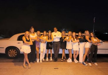 Group of friends and one man posing with arms crossed in front of a white stretch limousine at night, dressed in casual and dressy party outfits.