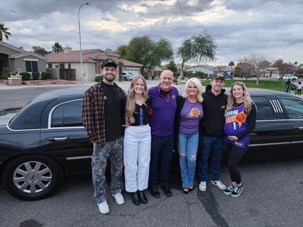 Six smiling adults wearing Phoenix Suns shirts and jerseys pose in front of a black stretch limousine on a suburban street under a cloudy evening sky.