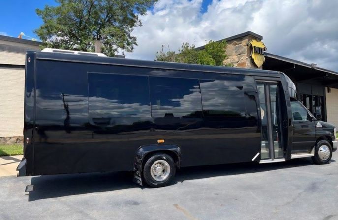 Glossy black shuttle/party bus with tinted windows and open side door parked outside a stone-and-brick building, clouds and blue sky reflected on its side.