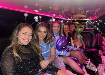 Five friends in sparkly and alien-themed costumes smiling and posing inside a pink-lit limousine, holding inflatable alien toys and party accessories.