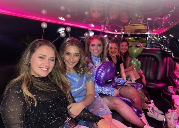 Five friends in sparkly and alien-themed costumes smiling and posing inside a pink-lit limousine, holding inflatable alien toys and party accessories.