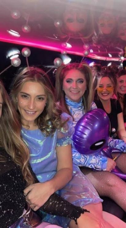 Five friends in sparkly and alien-themed costumes smiling and posing inside a pink-lit limousine, holding inflatable alien toys and party accessories.