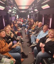 Large group of smiling adults seated facing each other inside a neon-lit party bus with pink and purple LED panels, enjoying a festive night out with drinks and snacks.