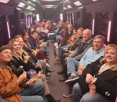 Large group of smiling adults seated facing each other inside a neon-lit party bus with pink and purple LED panels, enjoying a festive night out with drinks and snacks.