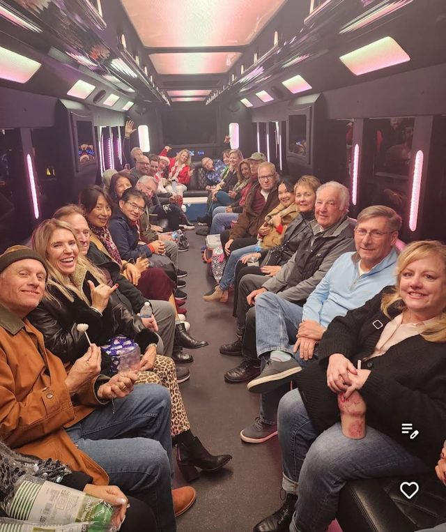 Large group of smiling adults seated facing each other inside a neon-lit party bus with pink and purple LED panels, enjoying a festive night out with drinks and snacks.
