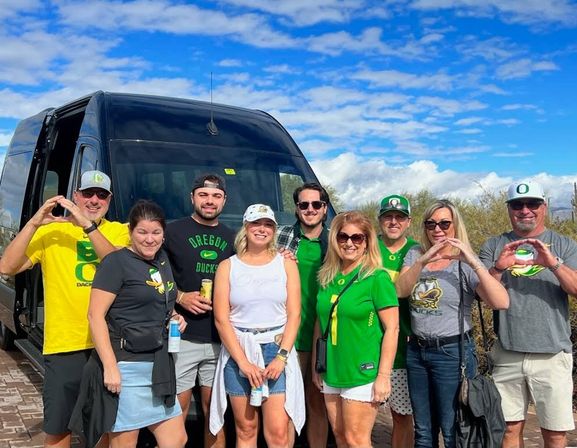 Smiling group of Oregon Ducks fans in green and yellow shirts and caps posing in front of a black passenger van under a bright blue sky — casual tailgate/road‑trip scene.