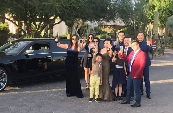 Well-dressed group of adults and children raising champagne glasses for a celebratory toast beside a black stretch limousine on a sunlit driveway with cacti and desert landscaping.
