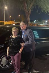 Woman in a sequined top and man in a blazer posing by a black stretch limousine in a lit parking lot at night.