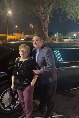 Woman in a sequined top and man in a blazer posing by a black stretch limousine in a lit parking lot at night.