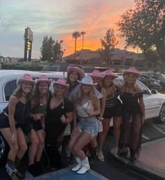 Group of eight women in party outfits and pink cowgirl hats smiling and posing by a white stretch limousine at sunset with palm trees and hills in the background
