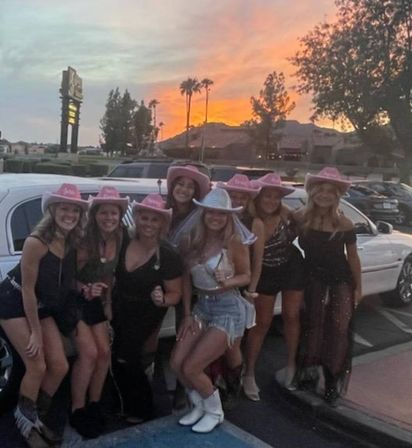 Group of eight women in party outfits and pink cowgirl hats smiling and posing by a white stretch limousine at sunset with palm trees and hills in the background
