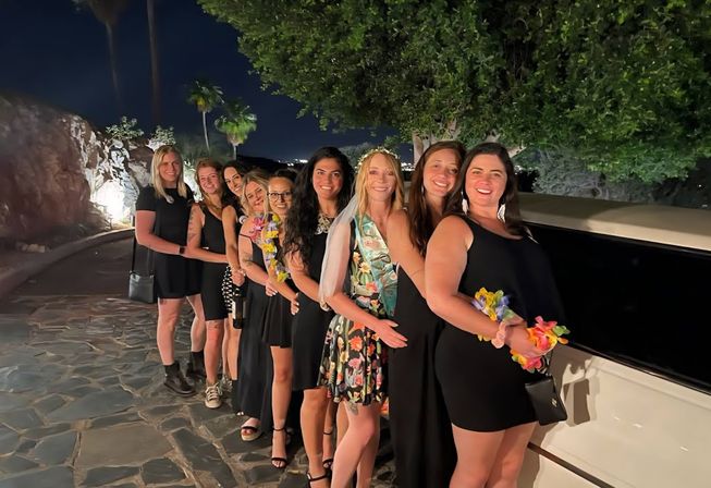 Outdoor nighttime bachelorette celebration: a group of women in black dresses and one in a floral sash and veil posing in a line by a white stretch limo on a palm‑tree lined stone driveway.