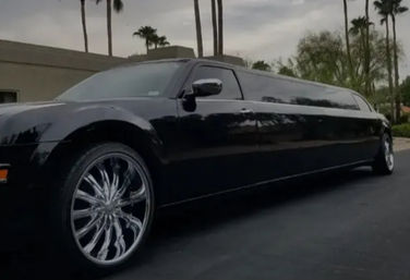 Glossy black stretch limousine with oversized chrome swirl rims parked on a palm-lined suburban street under a cloudy sky