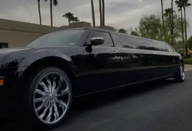 Glossy black stretch limousine with oversized chrome swirl rims parked on a palm-lined suburban street under a cloudy sky