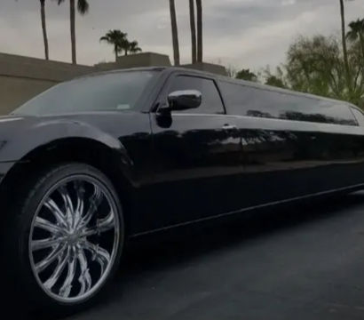 Glossy black stretch limousine with oversized chrome swirl rims parked on a palm-lined suburban street under a cloudy sky
