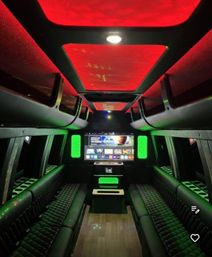 Luxury party bus interior with black quilted leather bench seating, wood plank floor, large flat-screen entertainment center and dramatic red ceiling panels with green LED accent lighting.