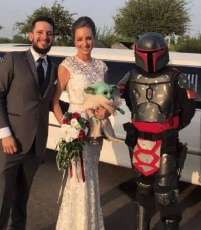 Bride in white lace gown and groom in gray suit smile beside a person in red-and-black Mandalorian armor while the bride holds a Baby Yoda (Grogu) plush in front of a white stretch limo at an outdoor wedding.