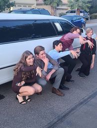 Five teenagers in formal outfits crouching and striking playful poses in front of a white stretch limousine on a suburban street with palm trees and parked cars