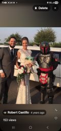 Bride in lace gown and groom in suit pose with a Mandalorian-style armored cosplayer holding a plush Grogu and a red-white bouquet in front of a white stretch limo outdoors.