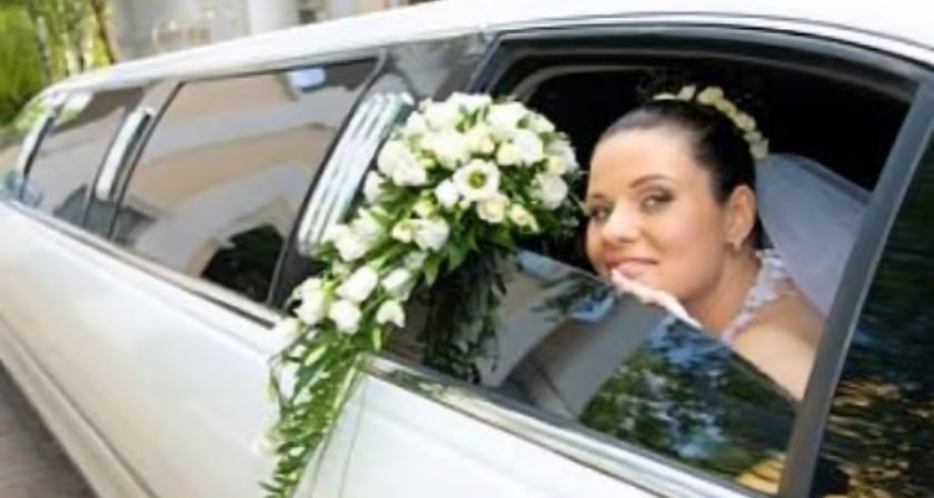 Smiling bride peeks from a white wedding limousine window holding a cascading white rose bouquet — bridal portrait in a wedding car.