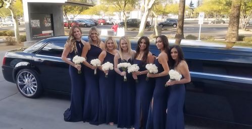 Seven bridesmaids in navy gowns holding white bouquets pose by a black stretch limousine in a sunny parking lot with palm trees