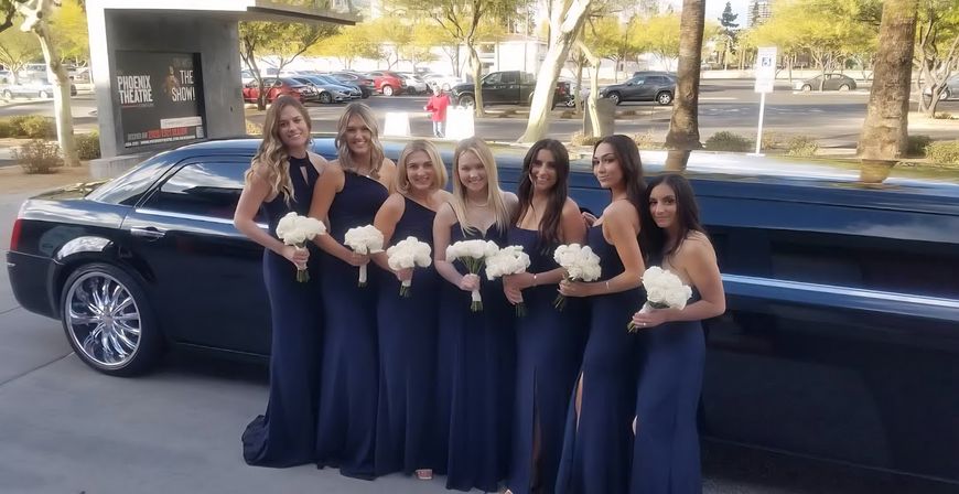 Seven bridesmaids in navy gowns holding white bouquets pose by a black stretch limousine in a sunny parking lot with palm trees