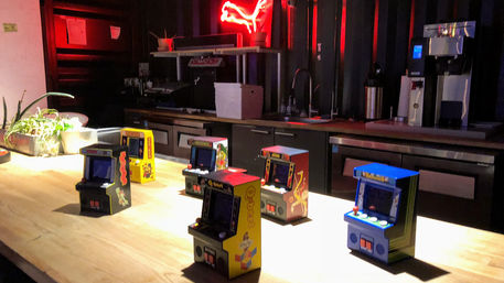 Row of colorful mini arcade cabinets on a long wooden countertop in a dimly lit indoor lounge/kitchen, with plants, coffee equipment and a red neon sign glowing in the background.