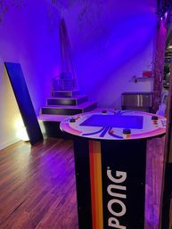 Purple-lit indoor game lounge with a retro Pong cocktail arcade table in the foreground, tiered black platform and vintage monitor on steps in the background, hardwood floor and moody ambient uplighting.