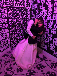 Bride in a white gown and partner in a suit dip for a kiss inside a neon-purple art-filled photo booth with bold black-and-white abstract mural and patterned floor