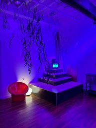 Purple-blue lit gallery interior with hanging foliage, tiered platform and vintage monitor, red modern chair and glowing orb on wooden floor