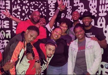 Group of friends posing and smiling in a purple-lit urban lounge, wearing casual jackets and hats against a bold black-and-white abstract mural — lively nightlife vibe.