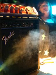 Live music stage close-up of a black guitar amplifier with a red effects unit on top, fog drifting across the speaker grille and a blurred musician wearing star-patterned clothing in colorful stage lights.
