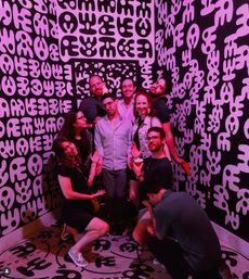 Group of friends striking playful poses in a neon-pink lit art installation with bold black-and-white abstract glyphs covering the walls and floor.
