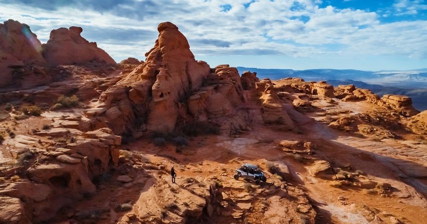 Aerial view of red-rock desert landscape with dramatic sandstone formations, a parked off-road UTV and a lone person under a blue sky with scattered clouds.
