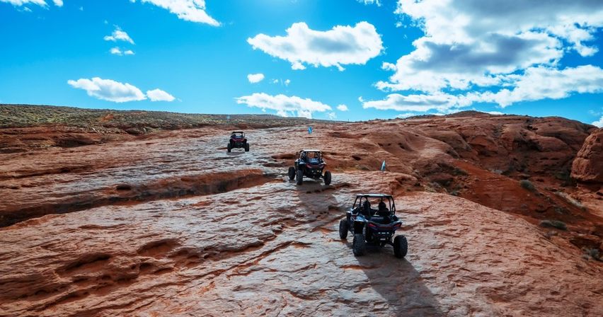 Three off-road UTVs driving across red sandstone slickrock in a sunlit desert landscape under a bright blue sky with scattered clouds.