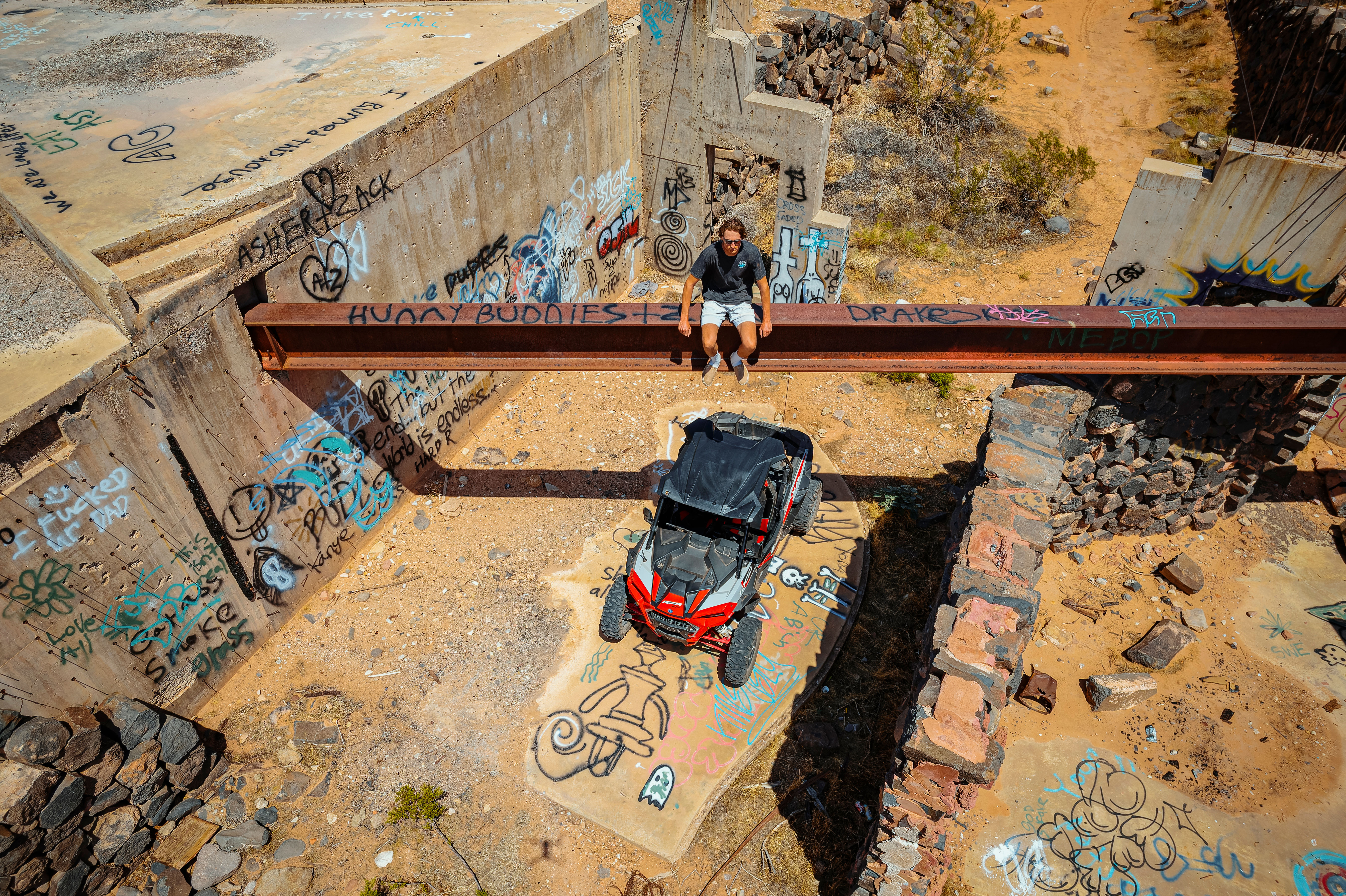 Aerial view of sunlit desert ruins covered in colorful graffiti, with a red off‑road side‑by‑side UTV parked on a painted concrete slab and a person sitting on a rusted steel beam above.
