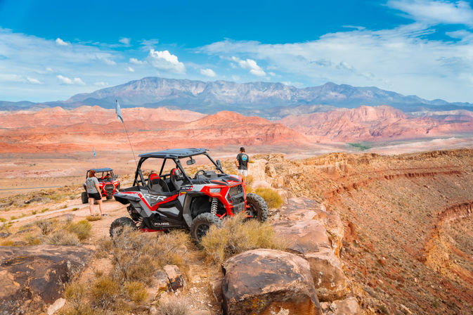 Two red-and-black off-road side-by-side UTVs parked on a rocky cliff in a red-rock Southwestern desert, overlooking a vast canyon and distant mountains under a bright blue sky with two people standing nearby.