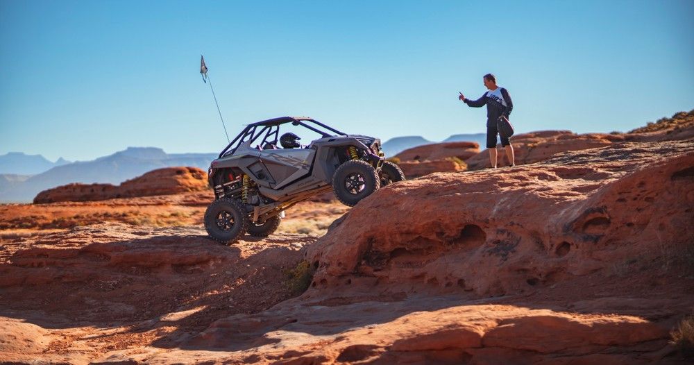 Off-road UTV climbing a red sandstone ledge in a red rock desert canyon while a person in off-road gear gives a thumbs-up under a clear blue sky