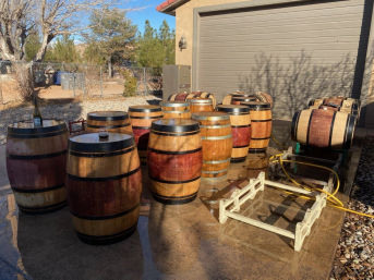 Sunlit backyard scene with wooden wine barrels arranged on a wet concrete driveway beside a closed garage, barrel racks, yellow hose and bare trees.