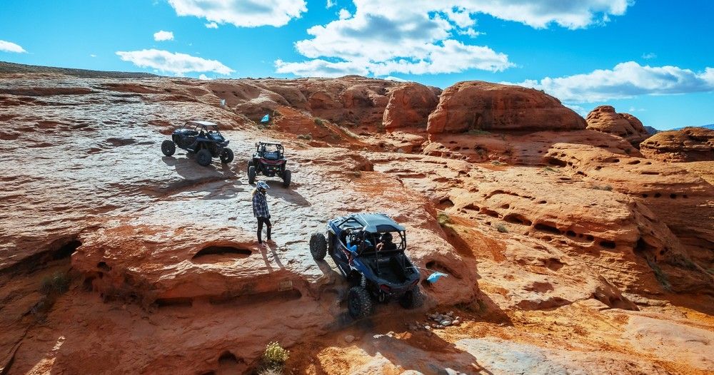 Three off-road UTVs and a person on red sandstone slickrock under a bright blue sky, navigating a red‑rock desert canyon — scenic off‑road adventure in the American Southwest.