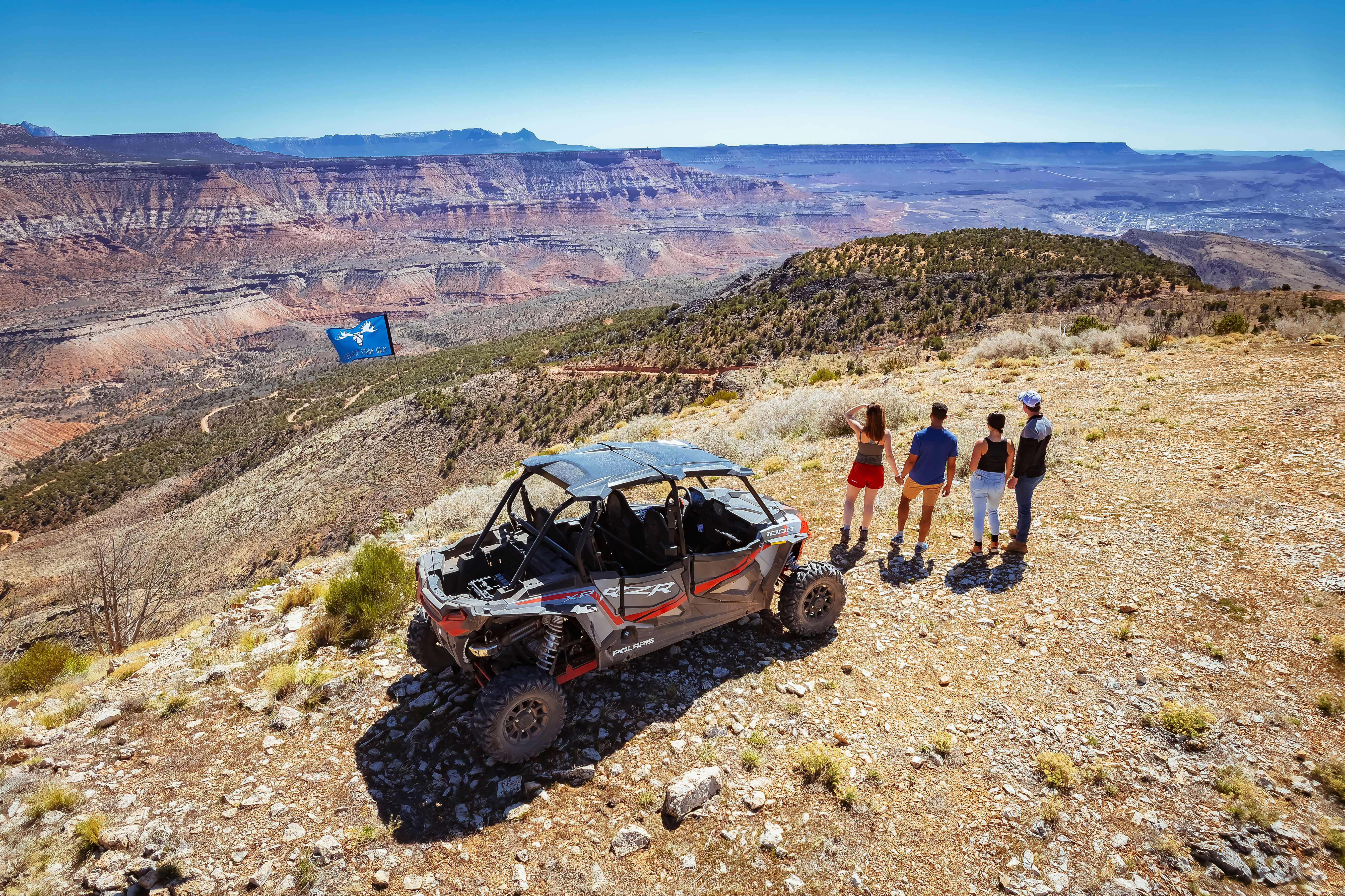 Group of four standing beside a parked off-road side-by-side UTV on a rocky plateau, overlooking a sweeping red-rock canyon and layered mesas under a bright blue sky.