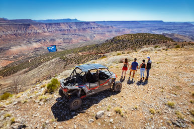 Group of four standing beside a parked off-road side-by-side UTV on a rocky plateau, overlooking a sweeping red-rock canyon and layered mesas under a bright blue sky.