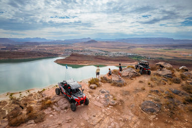 Red off-road UTVs parked on a rocky desert cliff as three people take in a scenic canyon lake overlook with turquoise water, sandy shorelines and distant mesas under a cloudy sky