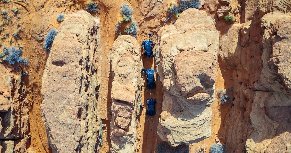 Aerial view of three off-road vehicles driving single-file through a narrow red-rock canyon in the Southwestern desert, surrounded by sandstone formations and sparse scrub.
