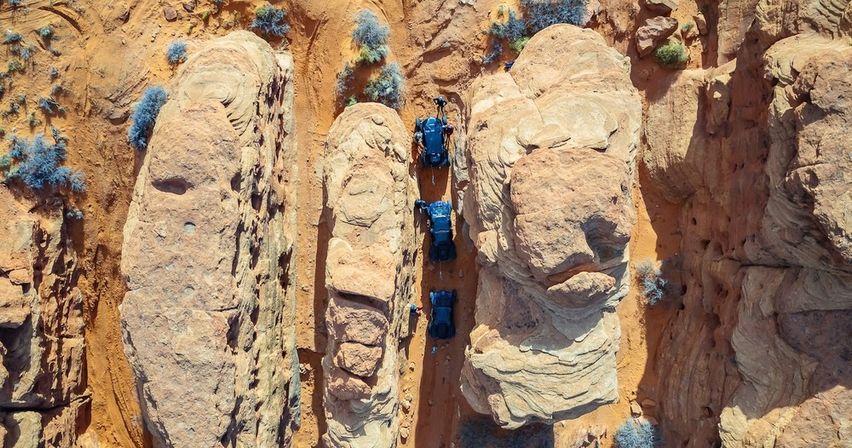Aerial view of three off-road vehicles driving single-file through a narrow red-rock canyon in the Southwestern desert, surrounded by sandstone formations and sparse scrub.