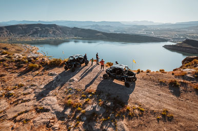 Two off-road UTVs and a small group on a rocky desert overlook, enjoying a panoramic canyon and lake view in golden-hour light.